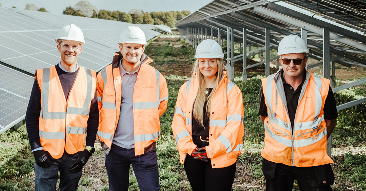A group photo of four individuals in safety gear stood smiling in front of a solar farm.